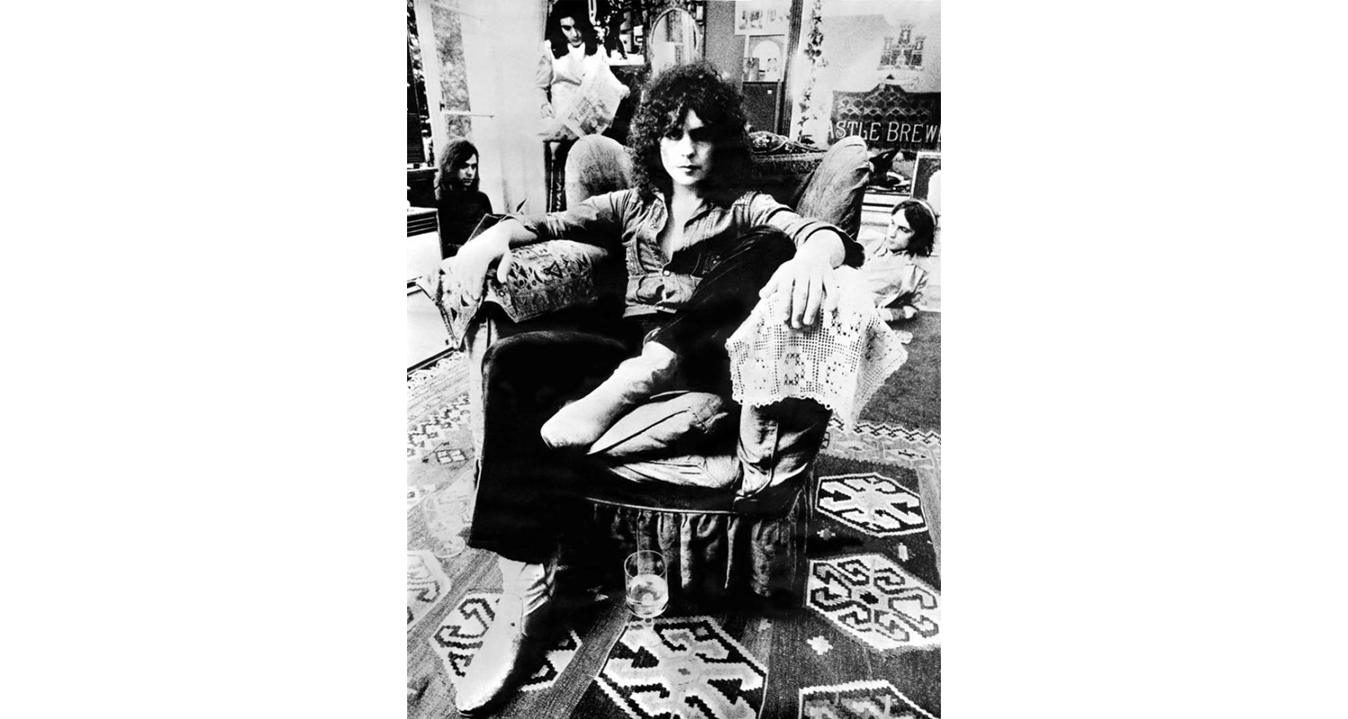 Rugs and Rockers: The Strange Bond Between Persian Rugs and Rock’n’Roll 18 Marc Bolan, frontman of psychedelic folk-rock band T. Rex, seated in his London home (c. 1970). The room's interior floor is decorated with geometric flatweave kilim rugs, photographed by Keith Morris.